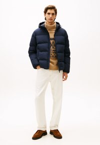 Giacca puffa blu navy con cappuccio, maglione lavorato a maglia beige con motivo, pantaloni crema e scarpe in suede marrone. La texture del tessuto è morbida e gonfia.