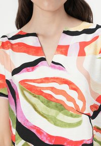 Bluză cu mânecă scurtă și decolteu în V, cu un model abstract îndrăzneț în roșu, verde, roz și negru pe un fundal alb. Textură fină a țesăturii.