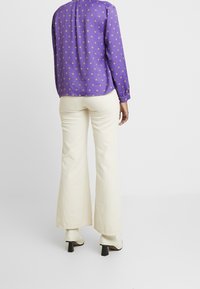 Blusa de manga larga morada con lunares dorados, combinada con pantalones acampanados color crema y botines de tacón color crema.