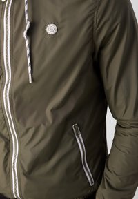 Olivgrüne Bomberjacke mit Reißverschluss vorne, kontrastierendem weißen Reißverschluss, Seitentasche und Logo-Patch auf der Brust. Glattes, leichtes Material.