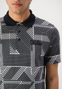 Camiseta polo a rayas en blanco y negro que presenta patrones geométricos, un cuello negro y un parche de logo en la zona del pecho.
