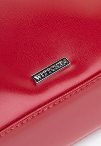 Czerwona torba z syntetycznej skóry o gładkiej fakturze, z metalowym logo marki w kolorze srebrnym. Szwy ozdobne wzdłuż krawędzi.