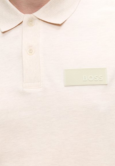 Gros plan d'un polo beige avec deux boutons et un patch rectangulaire avec le mot "BOSS" embossé sur la poitrine.