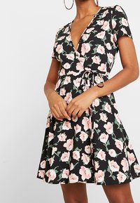 Kvinna som bär en svart blommig omslagsklänning med korta ärmar och knytning i midjan, accessoriserad med stora hoopsörhängen.