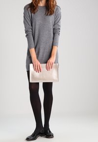 Abito maglione grigio in maglia con maniche lunghe, collant neri e scarpe nere lucide. Tiene in mano una pochette metallica argentata. Design semplice, senza motivi.