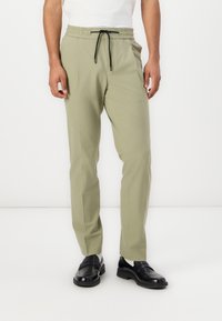 Light olive-green pants with an elastic waistband and black drawstring. Tailored fit with side pockets, paired with black loafers.