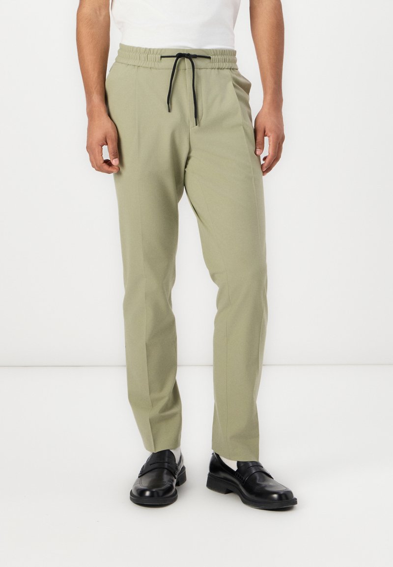 Helles olivgrünes Kleid mit elastischem Bund und schwarzem Zugband. Taillierte Passform mit Seitentaschen, kombiniert mit schwarzen Loafers.