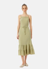Abito midi verde chiaro con spalline sottili, tessuto testurizzato, corpetto aderente e orlo a rouches a più strati. Indossato con scarpe beige a fantasia.