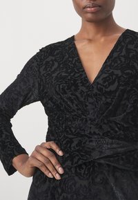 Robe en velours noir avec un décolleté en V et des manches longues. Motif floral texturé avec des plis à la taille. Texture lisse et douce.