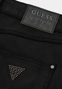 Jeans negros con etiqueta de cuero texturizado que dice "GUESS Los Ángeles EST. 1981". Un acento de triángulo con tachuelas metálicas en el bolsillo.