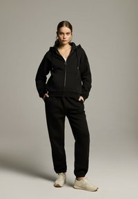 Czarna bluza z kapturem na zamek błyskawiczny, z regulowanym kapturem i przednimi kieszeniami, w połączeniu z pasującymi dresami. Wykonana z miękkiego materiału, o luźnym kroju.