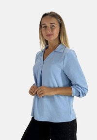Blouse bleu clair, à manches longues et col, texture douce et coupe décontractée. Dotée d'un décolleté en V et de poignets repliés, elle est associée à un pantalon noir.