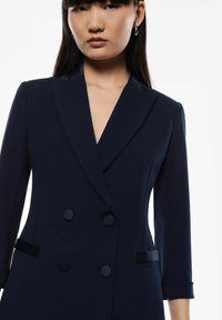 Blazer doppiopetto blu navy con revers, maniche a tre quarti e dettagli con bottoni; realizzato in un tessuto liscio e strutturato.