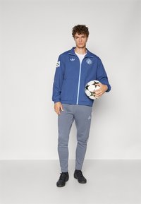 Blå lettvektsjakke med glidelås, hette og logoer; grå joggebukser med hvite striper; svarte fotballsko; holder en svart og hvit fotball.