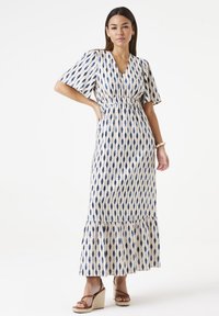 Maxikleid in Creme mit blauen, augenähnlichen Mustern, kurzen Puffärmeln und einem elastischen Tailleneinsatz. Es verfügt über einen volanartigen Saum und wird mit Wedge-Sandalen getragen.