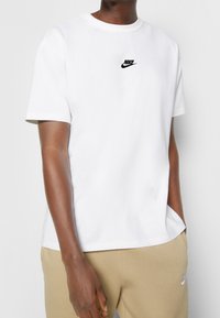 Bílé bavlněné tričko s kulatým výstřihem a krátkými rukávy. Na hrudi se nachází malé černé logo Nike. Hladký materiál, volný střih.