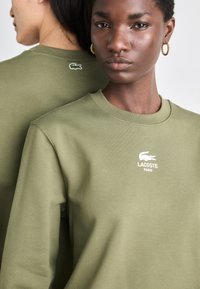 Olive zelená mikina vyrobená z mäkkej zmesi bavlny, s okrúhlym výstrihom, rebrovanými manžetami a predným logom "LACOSTE PARIS" v bielej farbe.
