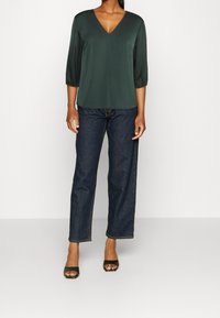 Grünes V-Ausschnitt-Bluse mit Dreiviertelärmeln, kombiniert mit dunkelblauen Jeanshosen und schwarzen Peeptoe-Absätzen. Glatte Stofftextur.