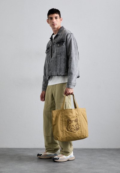 Tote bag de culoare căpru deschis cu un grafic de cap de lup, asortată cu o jachetă din denim gri, un tricou alb, pantaloni cargo verzi și adidași bej.