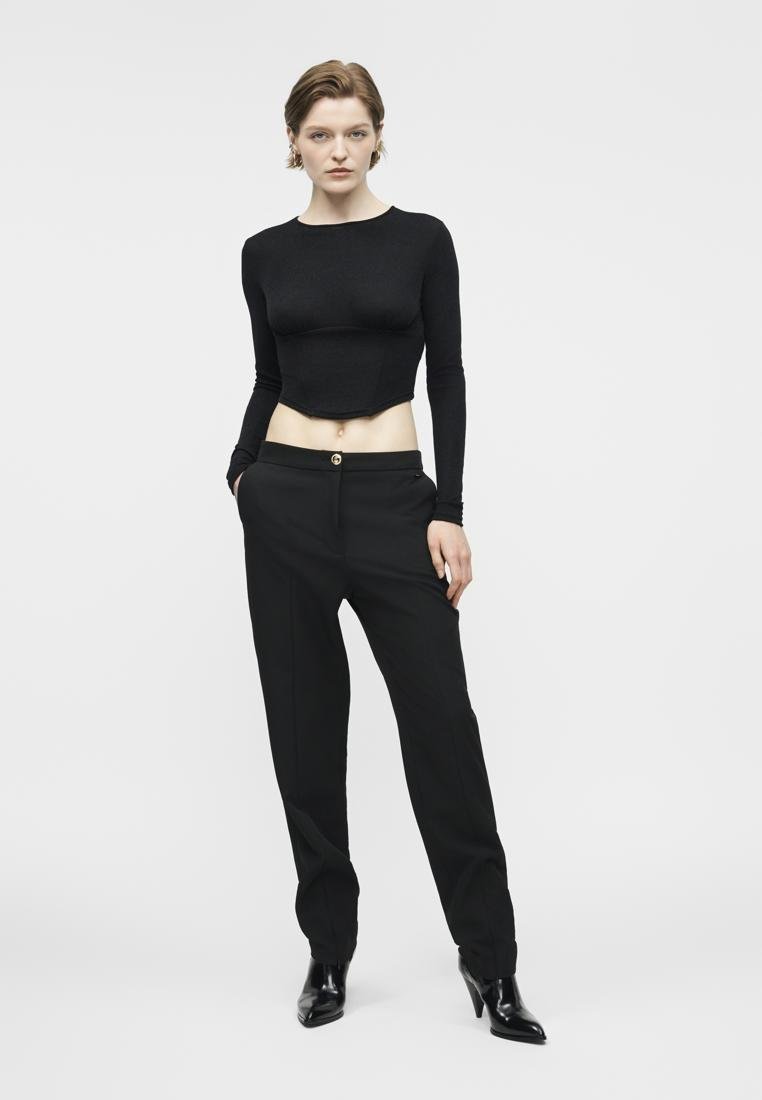 Jeune personne debout, mains dans les poches, portant un haut noir à manches longues raccourcies, un pantalon noir et des bottines noires à talons sur fond blanc.