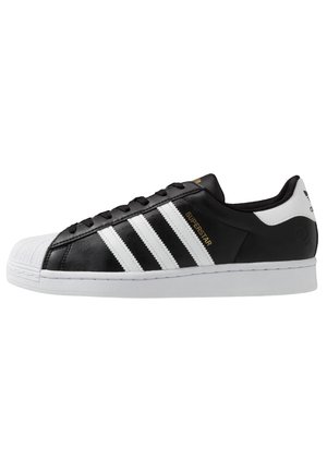 Baskets Adidas Superstar noir et blanc avec bout coquille blanc, trois bandes blanches et texte doré "Superstar" sur le côté.