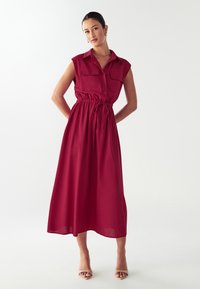 Abito midi senza maniche di colore bordeaux con colletto, due tasche sul petto, vita elasticizzata e gonna fluida. Tessuto dalla consistenza morbida.