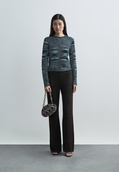 Mulher em pé vestindo uma blusa de mangas longas listrada, calças largas pretas com padrão, segurando uma bolsa com corrente multicolorida e sandálias de salto aberto.