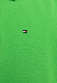 Zielona koszulka polo wykonana z teksturowanej tkaniny, z małym haftowanym logo w kolorach czerwonym, białym i niebieskim umieszczonym na lewej piersi.