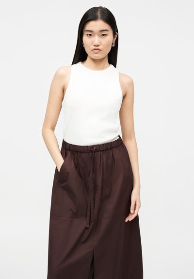 Mladá žena s dlhými tmavými vlasmi má na sebe biely top bez rukávov a tmavohnedú sukňu s vysokým pásom a sťahovacou šnúrkou, ruky voľne pri bokoch.