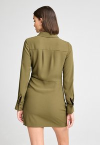 Olivengrünes Kleid mit Kragen, langen Ärmeln, knöpfbarem Verschluss vorne und Falten am Rücken, aus glattem, leichtem Stoff.