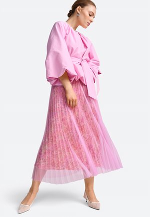 Frau trägt ein rosa Oberteil mit Puffärmeln und gebundener Taille sowie einen rosa plissierten Blumenrock, kombiniert mit hellen Slingback-Absatzschuhen.