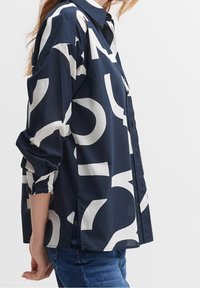 Chemise bleu marine à boutons avec des motifs abstraits blancs, manches longues avec des poignets retroussés et fentes latérales pour plus de liberté de mouvement. Tissu en coton.