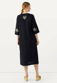 Schwarzes Leinenkleid mit Dreiviertelärmeln, verziert mit goldenen Stickereien an den Ärmeln und im Rückenteil, gerader Schnitt und seitlichen Schlitzen für mehr Bewegungsfreiheit.