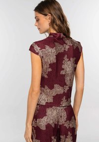 Top burdeos con un acabado texturizado, que presenta un patrón abstracto en dorado y un diseño de cuello simulado. Mangas principales y una silueta ajustada.