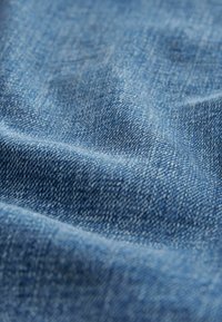 Denim tkanina srednje plave boje, s glatkom teksturom i suptilnim dijagonalnim tkanjem, prikazuje varijacije svjetla i sjene.