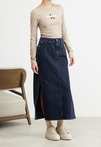 Marinblå denim maxikjol med en frontslits, parat med en beige långärmad topp med en logotyp, och krämfärgade sneakers. Trästol i bakgrunden.