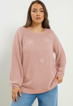 Kvinde iført en løs lyserød strikket sweater med 3D blomstermønstre, kombineret med lyseblå jeans, stående foran en ensfarvet baggrund.