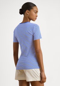 Γυναίκα με τα μαλλιά της σε κότσο, που φοράει χρυσούς κρίκους στα αυτιά, μια μπλε και άσπρη ριγέ κοντομάνικη μπλούζα και μπεζ σορτς, στραμμένη ελαφρώς στο πλάι.