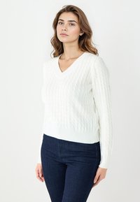 White knitted sweater with a V-neck, featuring a textured cable pattern and ribbed cuffs and hem, paired with dark blue jeans.