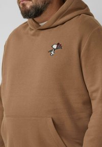 Brązowa bluza z kapturem wykonana z miękkiego materiału. Ozdobiona małym wyszywanym wzorem Snoopiego trzymającego patyk z kwiatem. Posiada przednią kieszeń.