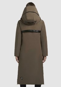 Parka lunga marrone con un ampio cappuccio, caratterizzata da una banda con logo nero sul retro e tasche laterali, realizzata in un materiale liscio e strutturato.
