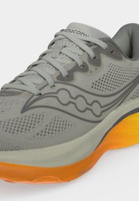 Γκρι παπούτσι τρεξίματος Saucony με πάνω μέρος από δίχτυ, γκρι κορδόνια και φωτεινή πορτοκαλί σόλα, που δείχνει το λογότυπο στο πλάι και μαλακή επένδυση στον κολάρο.