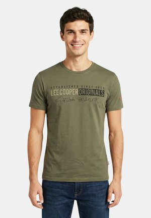 Joven que lleva una camiseta verde oliva de Lee Cooper Originals y vaqueros azules, sonriendo y de pie frente a un fondo blanco liso.
