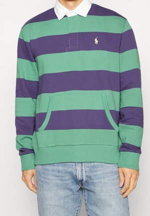 Homme portant un maillot de rugby rayé vert et violet avec un col blanc et une poche avant, associé à un jean bleu clair.