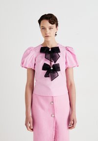 Kvinne iført en rosa topp med puffete ermer, med to store svarte sløyfer, og en matchende rosa knelang jakke mot en enkel hvit bakgrunn.