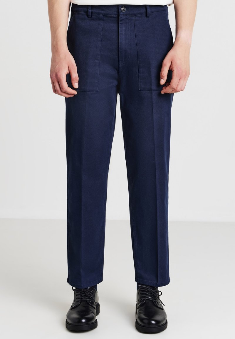Marineblaue Hosen mit schmaler Passform, aus glattem Stoff, mit zwei Vordertaschen und einem Knopfverschluss. Getragen mit schwarzen Schuhen.