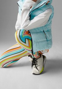 Světle modrá péřová vesta přes bílou mikinu, doplněná o vícebarevné pruhované legíny a bílé tenisky s černými tkaničkami a zelenými detaily.
