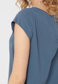 Blusa blu a maniche corte con tessuto texturizzato, caratterizzata da un colletto rotondo e un accento di catena argentata sul retro.