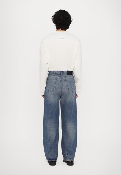 Weißes langärmliges Hemd mit einem Etikett auf der Rückseite, kombiniert mit locker sitzenden blauen Jeans, die ein schwarzes Etikett und einen ausgebleichten Look aufweisen.