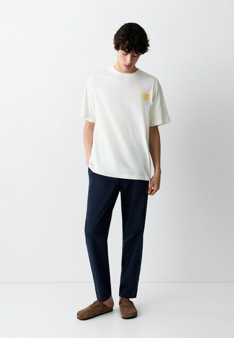 Jeune homme portant un t-shirt blanc « Good Times », un pantalon bleu marine et des chaussures marron sans lacets, debout devant un fond blanc uni.